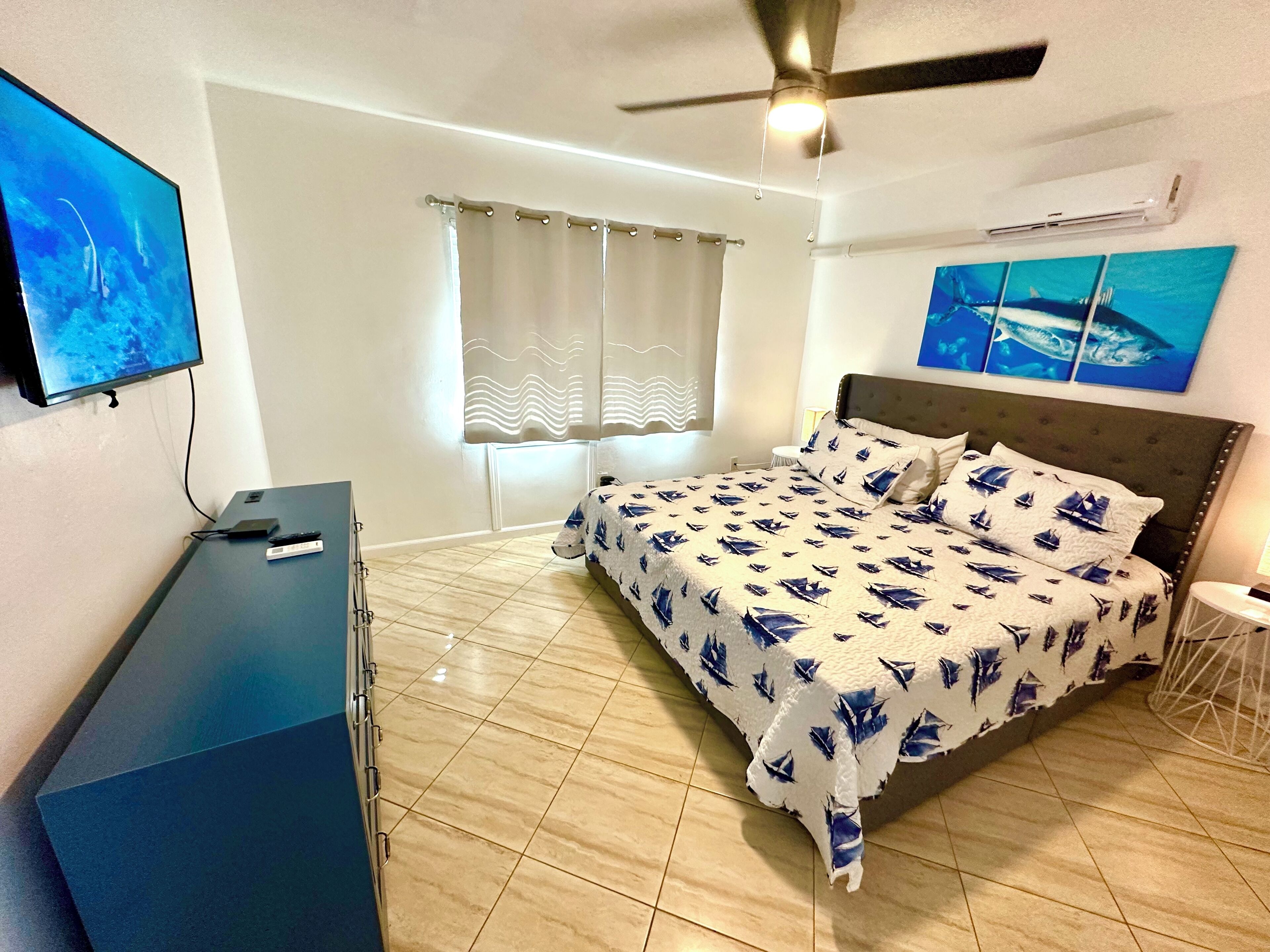 A bedroom with a bed, a desk, a TV, a ceiling fan, and artwork on the walls.