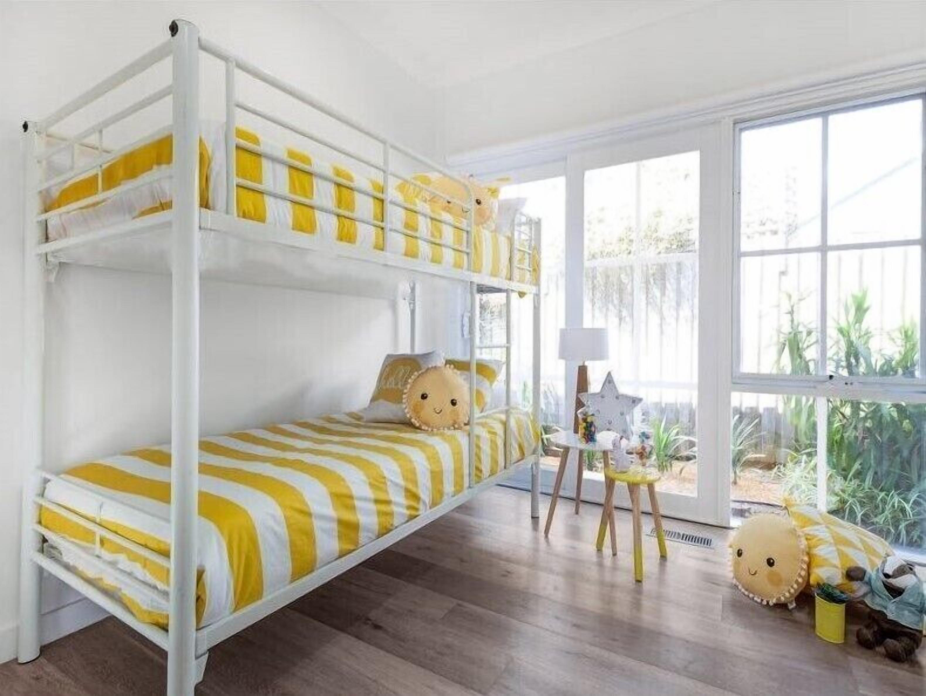 A bunk bed with yellow and white striped bedding, a small table with a lamp, and a window with a view of greenery.