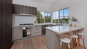 A modern kitchen with a central island, wooden flooring, and large windows offering a view of greenery. - Relaxed Sorrento Coastal Living (Sorrento)