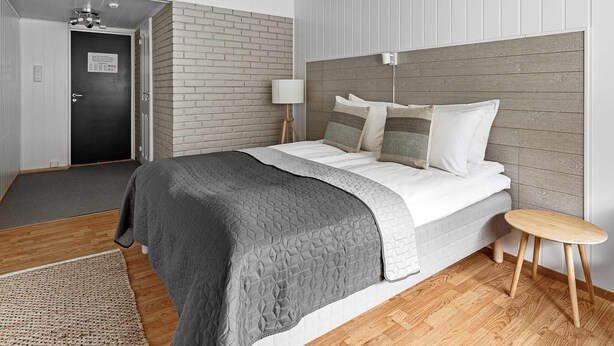 A modern bedroom with a brick-patterned headboard, a bed with a grey quilt, a small wooden stool, and a white lamp. - Eidfjord Hotel (Eidfjord)