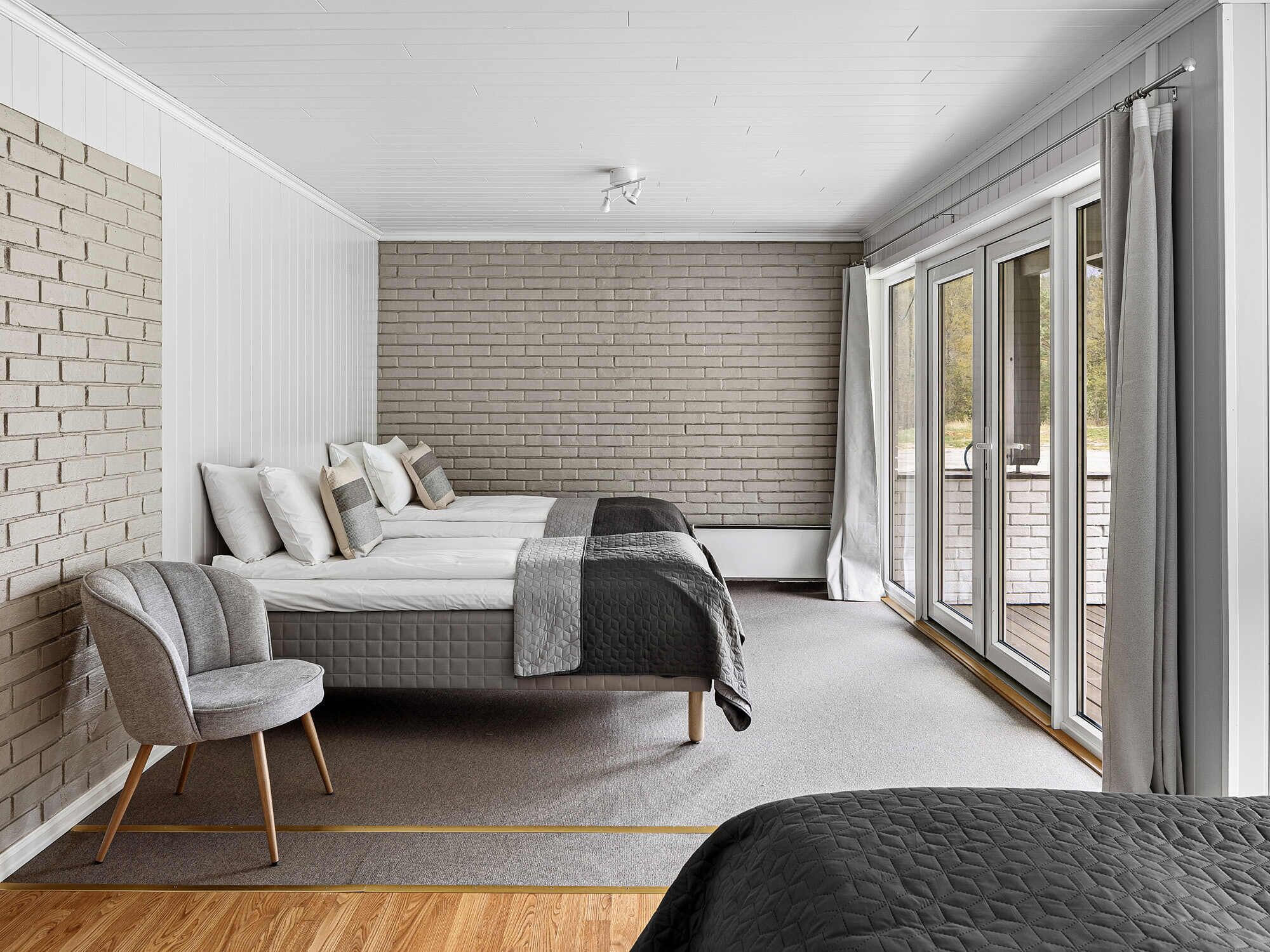 A bedroom with a brick wall, a bed with a grey quilt, a grey armchair, and large windows.