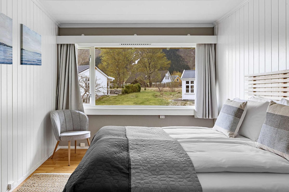 A bedroom with a large bed, a chair, and a window with a view of houses and trees.
