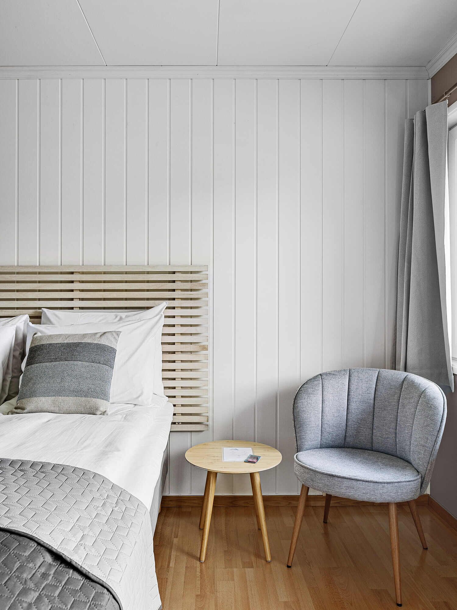 A modern bedroom with a wooden headboard, a grey upholstered armchair, a round wooden side table, and a bed with white bedding and a striped pillow.