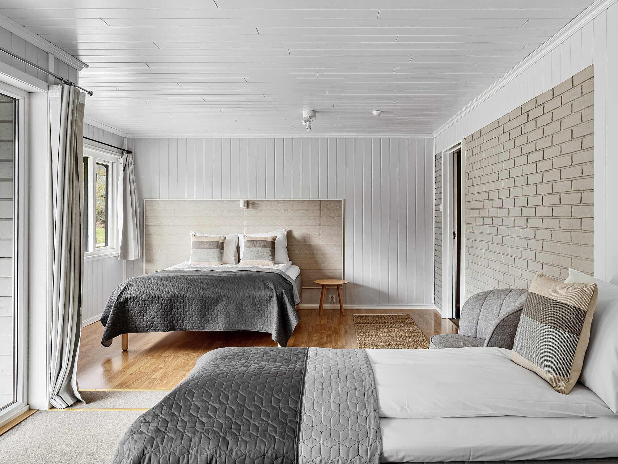 A modern bedroom with a large bed, a smaller bed, and a daybed. The room has a wooden ceiling, white walls, and a brick accent wall.