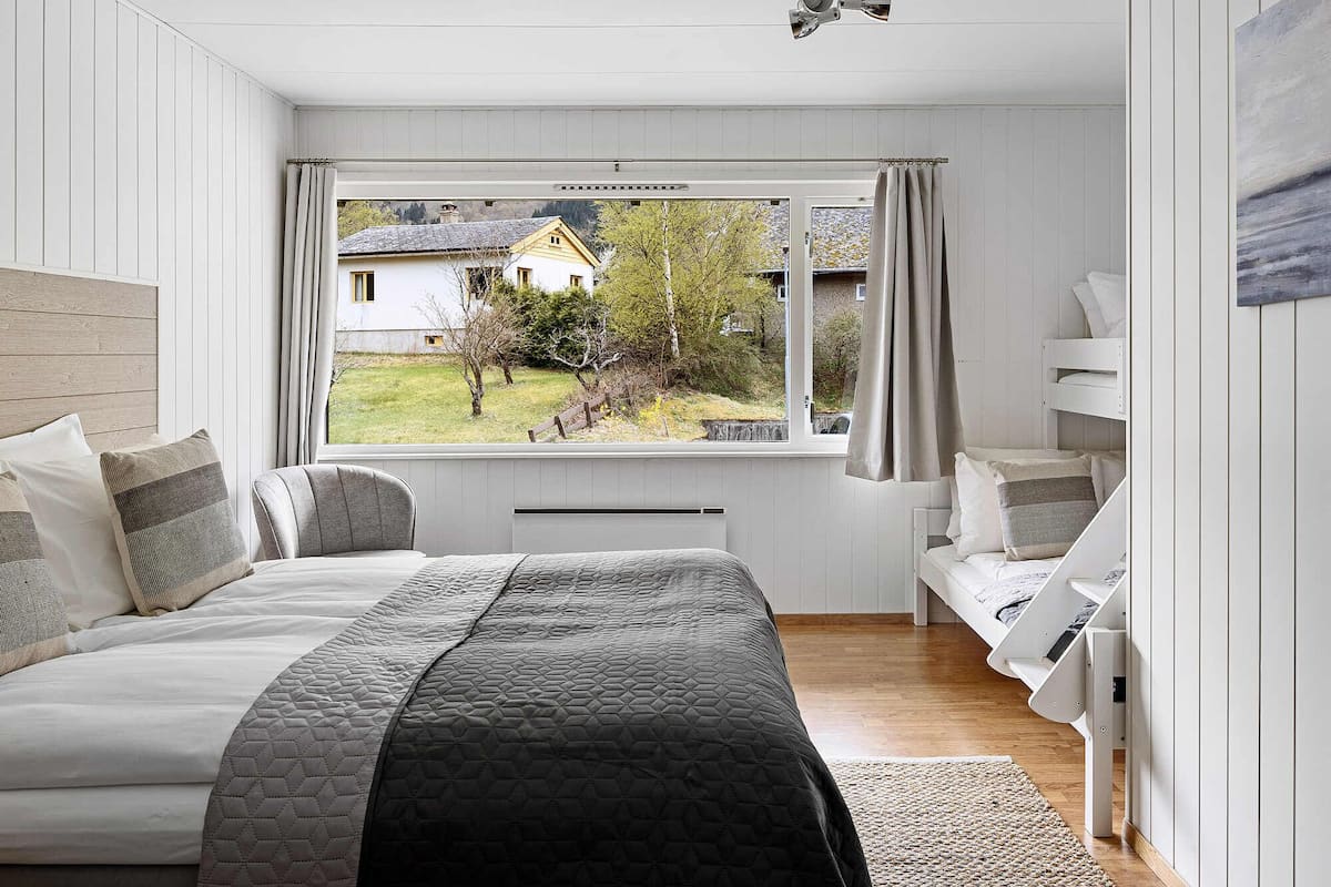 A bedroom with a large bed, a window with curtains, a ceiling fan, and a view of a house and trees outside.