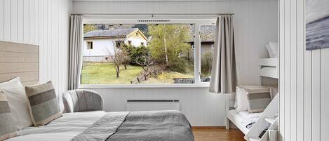 A bedroom with a large bed, a window with curtains, a ceiling fan, and a view of a house and trees outside.