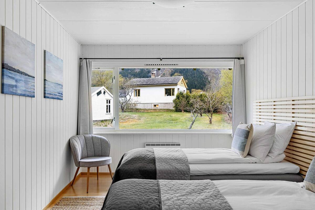 A bedroom with a large window, a bed, a chair, and a view of a house and trees.