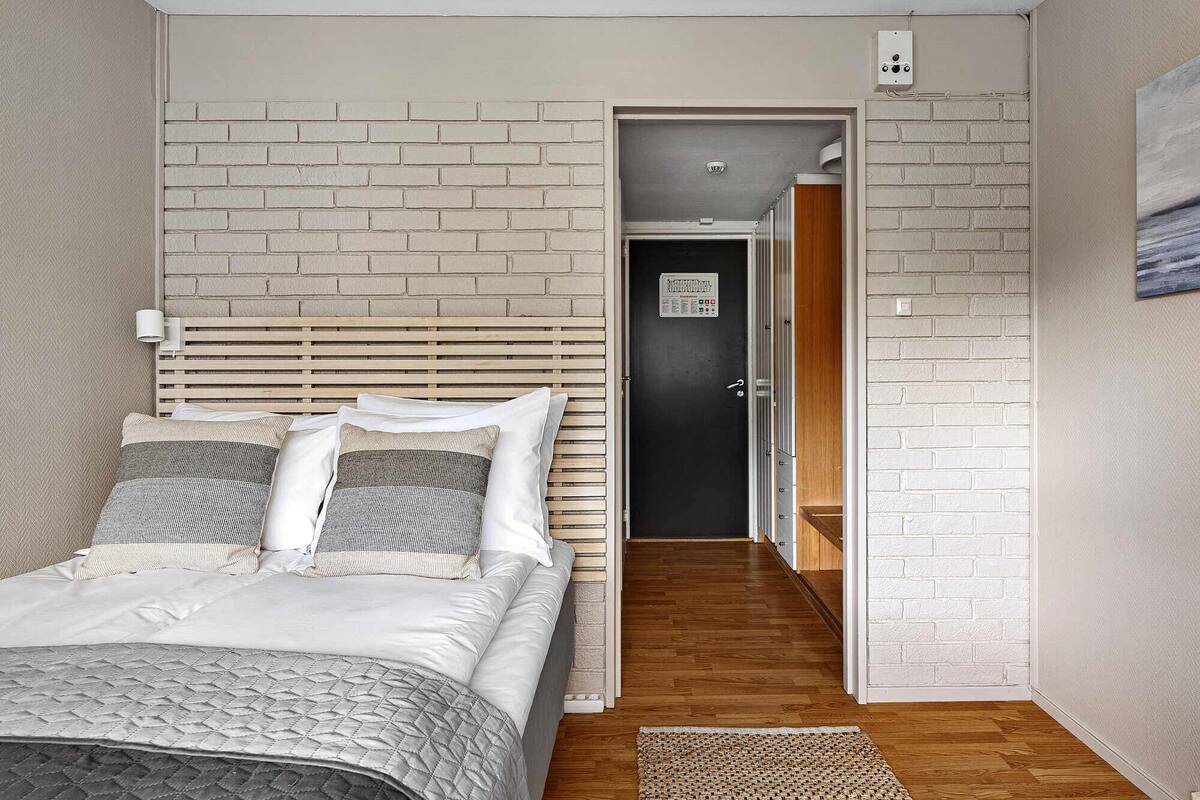 A bedroom with a bed, a wooden headboard, a brick-patterned wall, a door, and a hallway leading to another room.