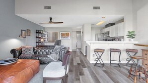 Una sala de estar moderna con un sofá de cuero, una zona de comedor con taburetes y una cocina con armarios blancos.