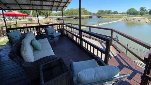 A covered deck with outdoor furniture, a view of a river, and a bridge in the background. - Cozy riverside cabin on the Neosho river (Chetopa)