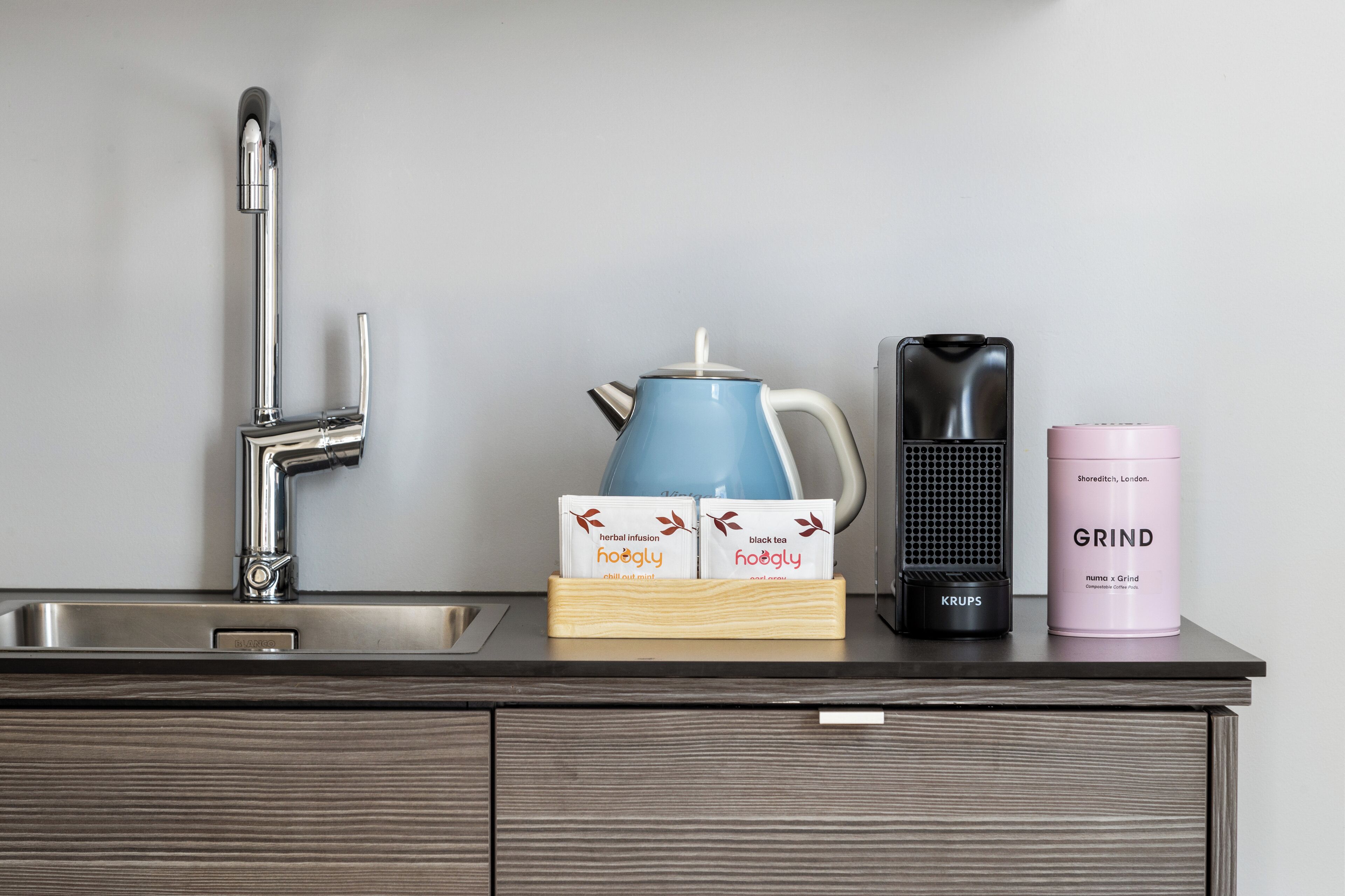 Un plan de travail de cuisine avec un évier, une bouilloire bleue, une machine à café noire et un récipient rose portant l’inscription « GRIND ».