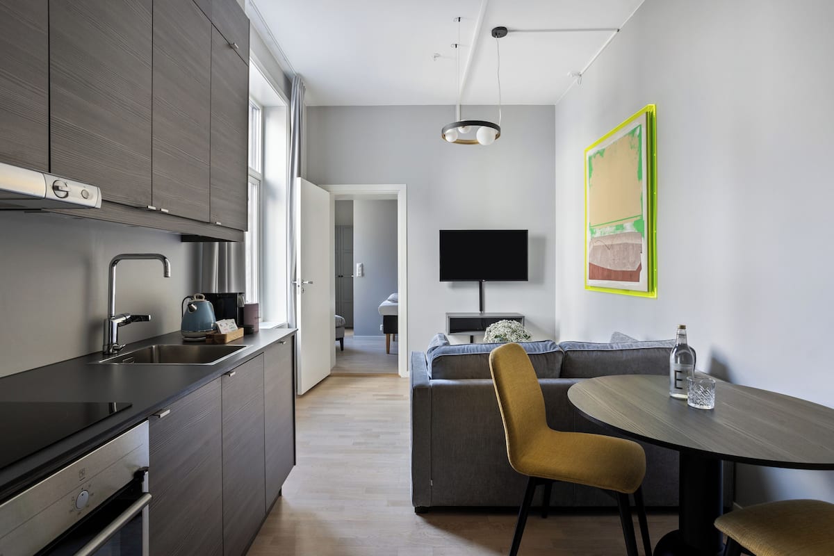 A modern kitchen with dark cabinetry, a stainless steel sink, and a built-in oven. There is a dining area with a round table and two chairs, and a flat-screen TV mounted on the wall.