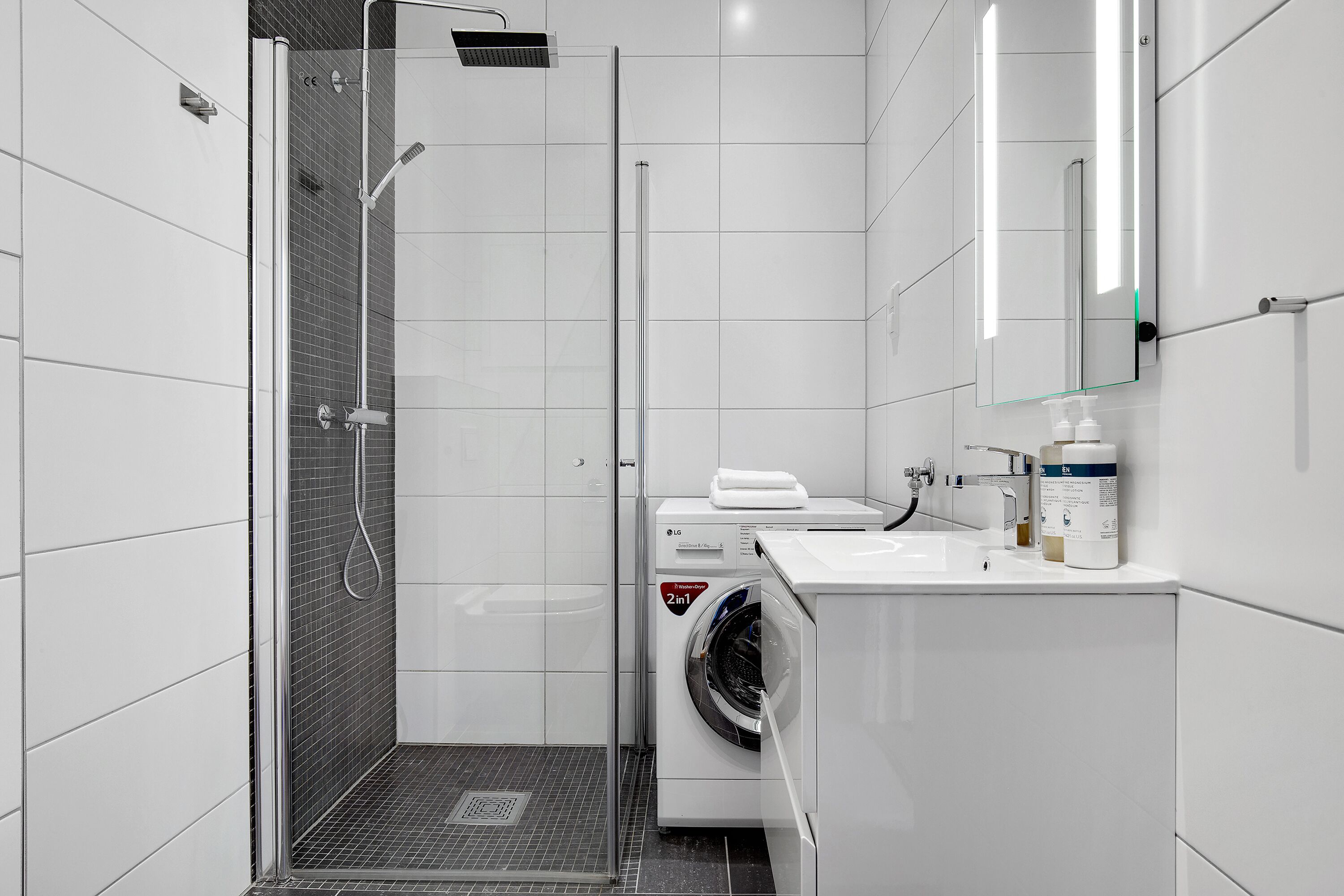Un baño moderno con ducha envuelta en vidrio, lavadora y lavamanos con espejo arriba.