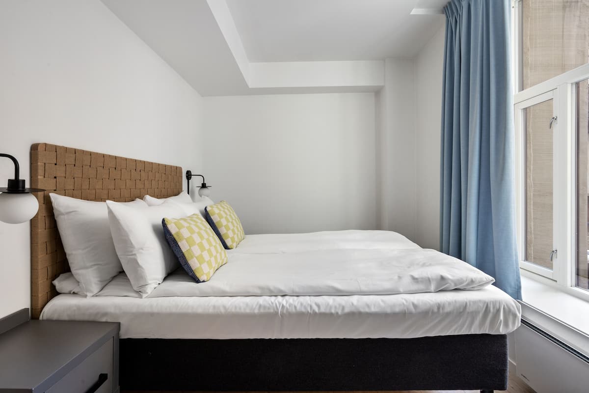 A neatly made bed with white bedding and two pillows, a wooden headboard, a bedside table with a lamp, and a window with blue curtains.