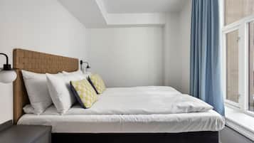 A neatly made bed with white bedding and two pillows, a wooden headboard, a bedside table with a lamp, and a window with blue curtains.