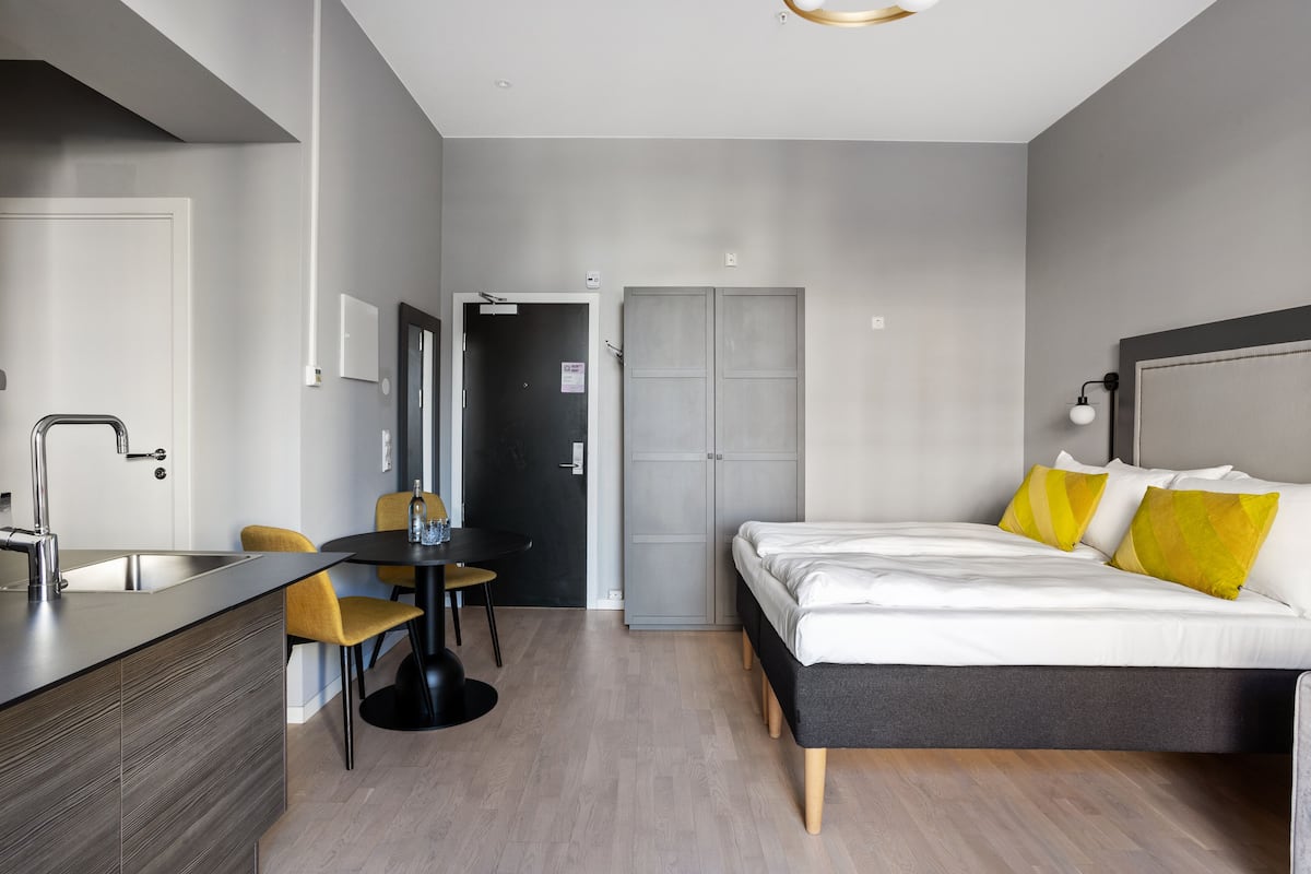 A modern hotel room with a bed, bedside table, and a small kitchenette.