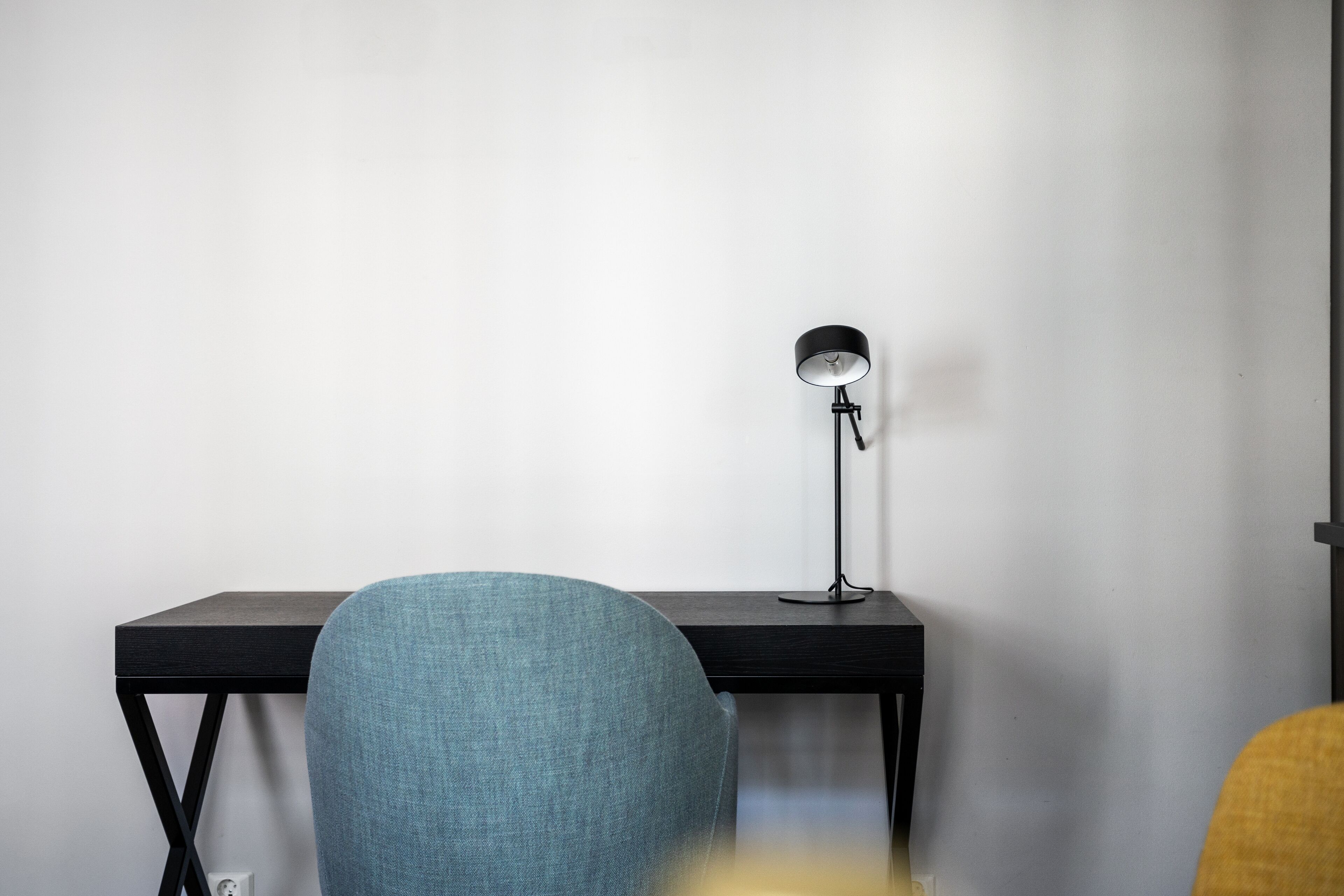 A modern workspace with a black desk, a blue chair, and a black floor lamp against a plain white wall.
