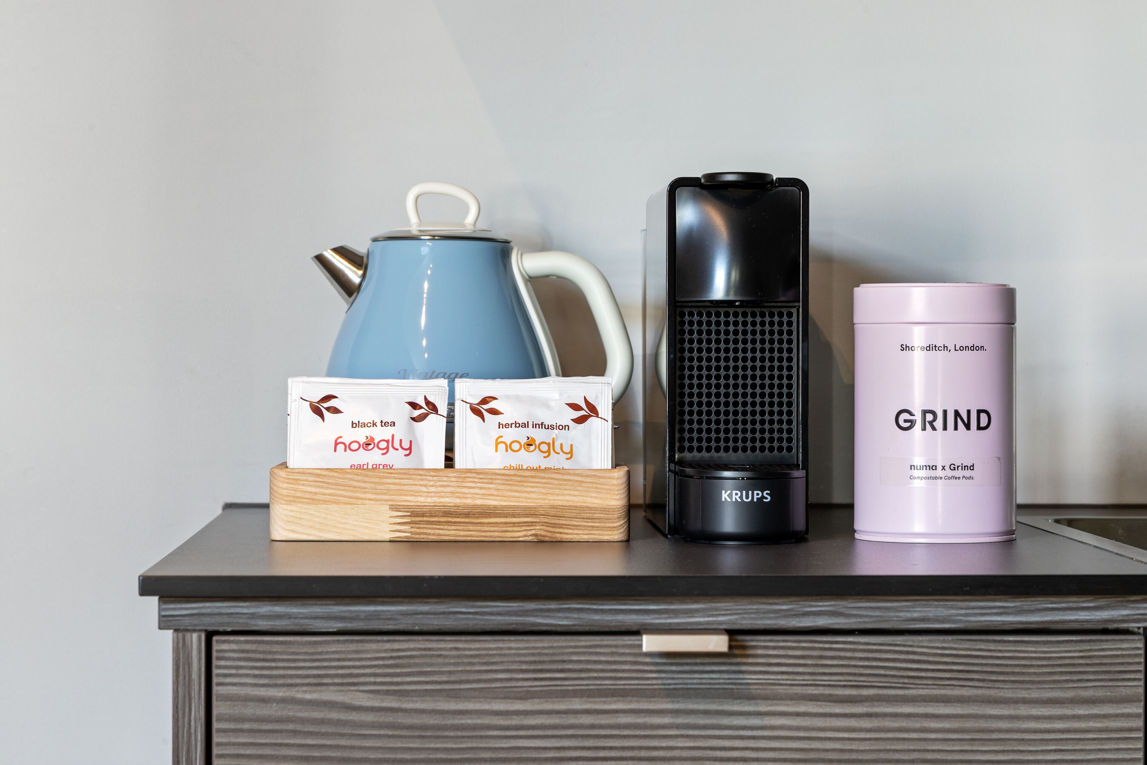 Un plan de travail de cuisine avec une bouilloire bleue, une machine à café noire et un récipient rose portant l’inscription « GRIND ».