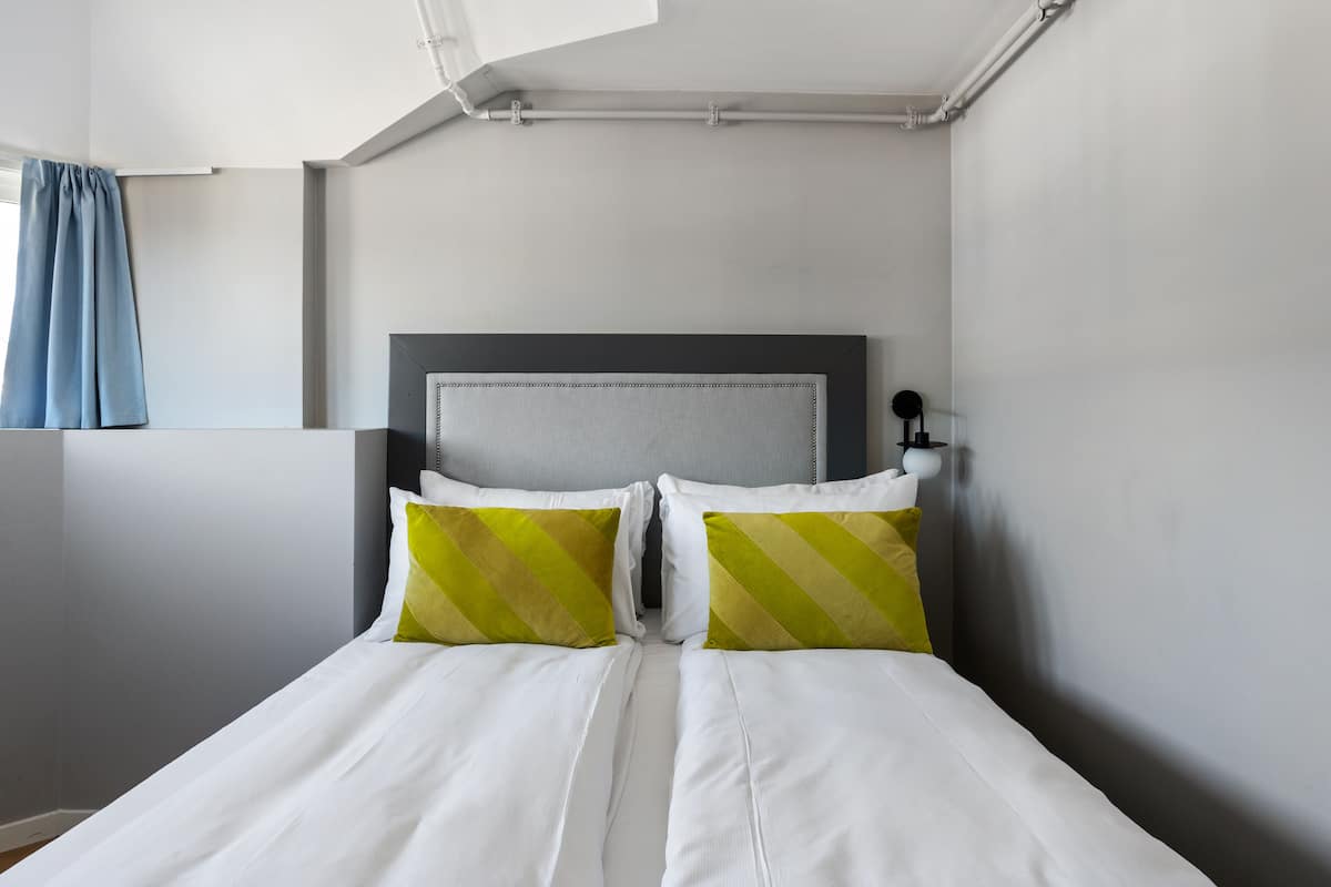 A neatly made bed with white and green striped pillows, a grey headboard, and a white wall with a window and curtain.