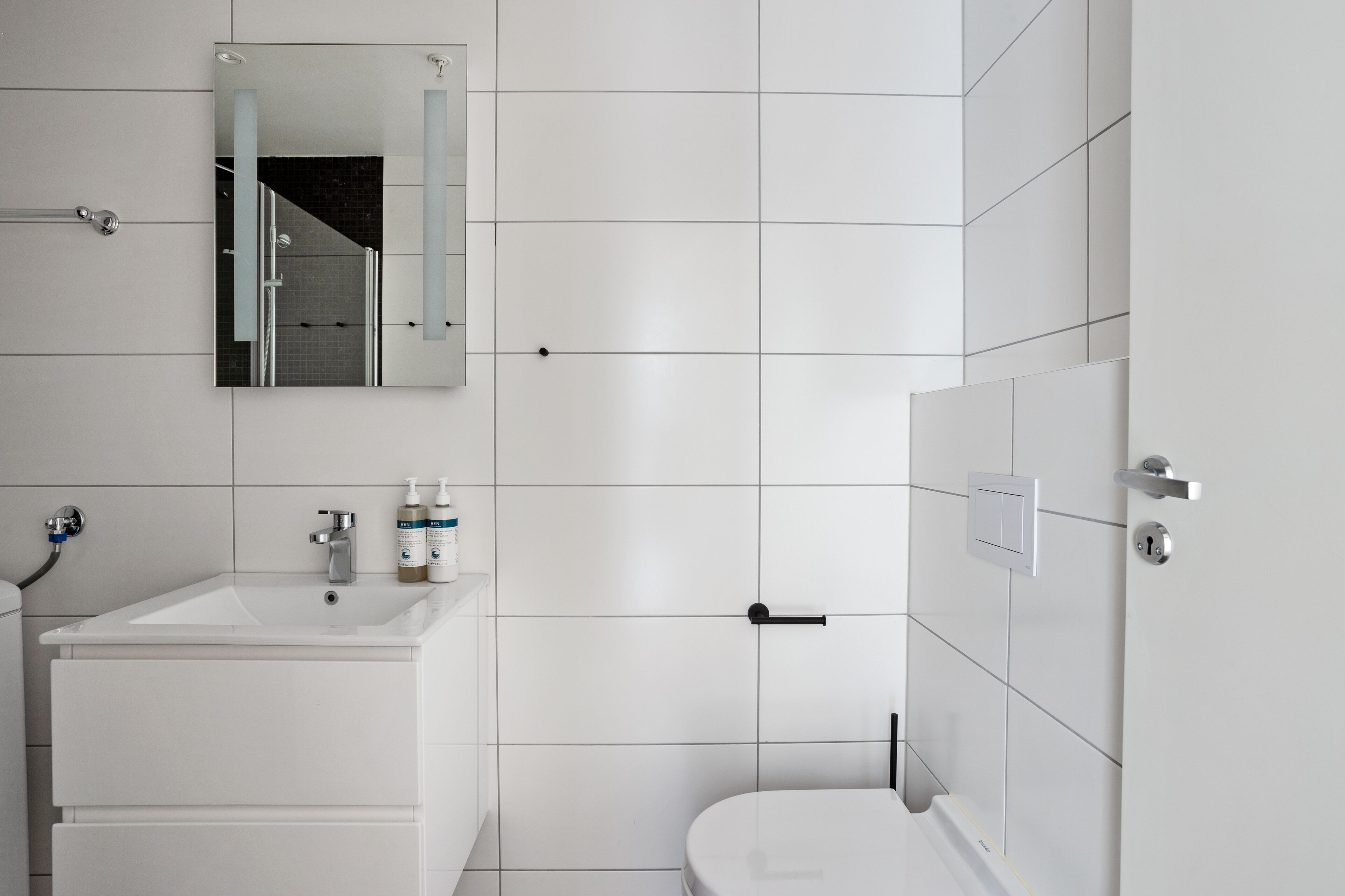 Une salle de bain moderne avec des carreaux blancs, un lavabo, des toilettes et un miroir.