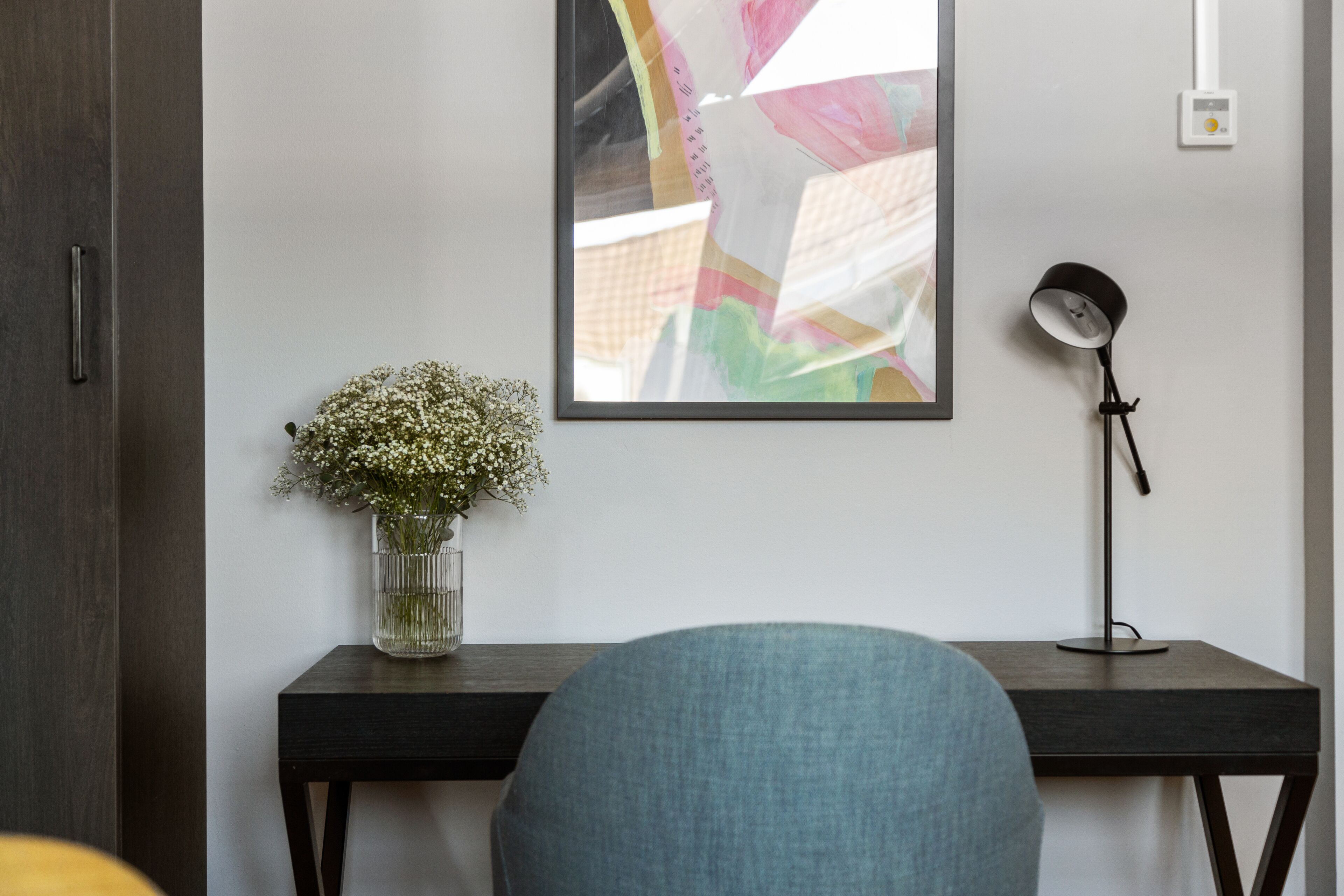 Une pièce moderne avec un bureau en bois foncé, un vase de fleurs, un tableau abstrait encadré et une lampe sur pied.
