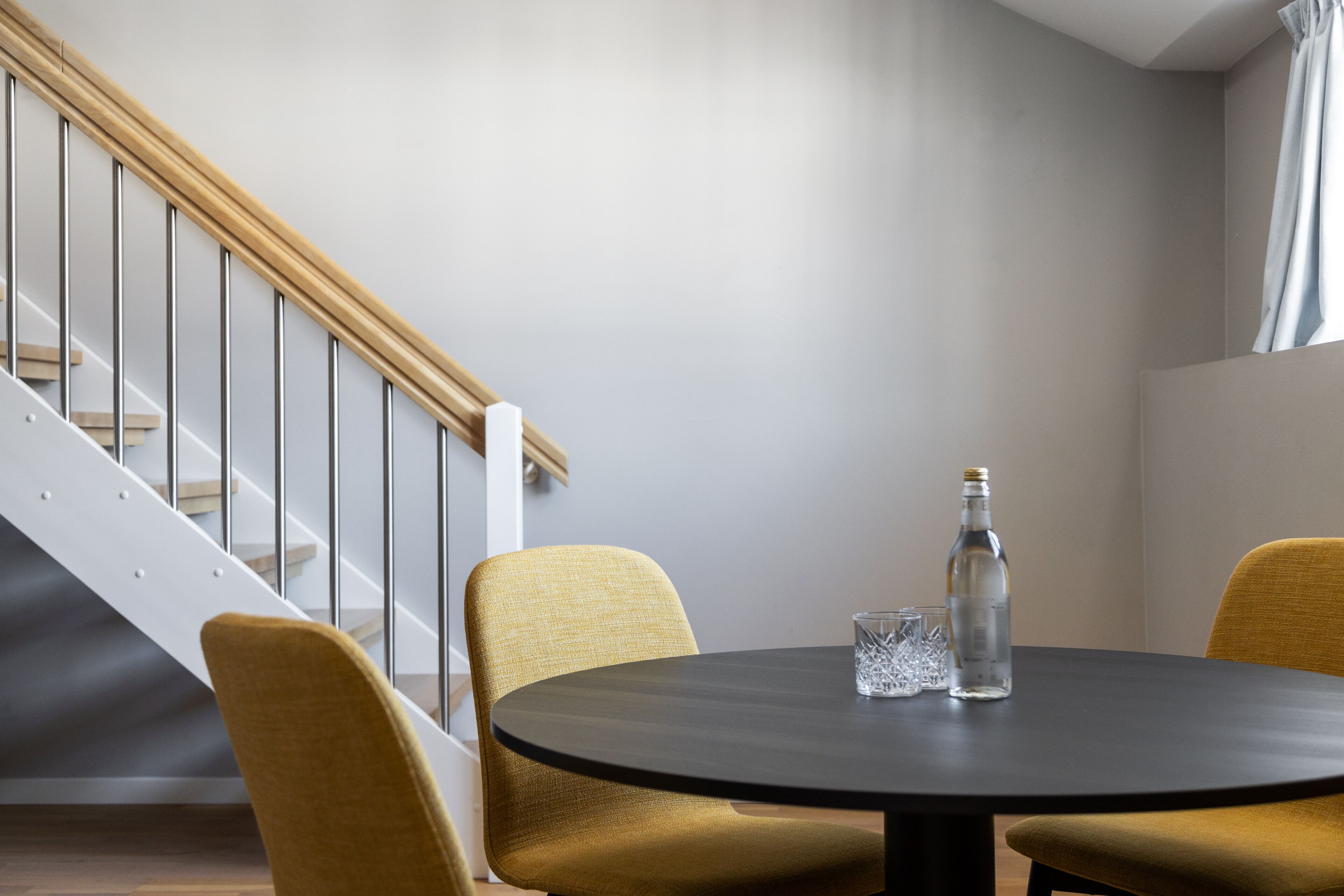 Una habitación moderna con una mesa redonda, dos sillas color mostaza, una botella de agua y un vaso sobre la mesa. Se observa una escalera con barandilla de madera y balaustradas de metal al fondo.