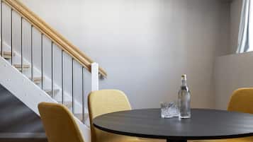 Una habitación moderna con una mesa redonda, dos sillas color mostaza, una botella de agua y un vaso sobre la mesa. Se observa una escalera con barandilla de madera y balaustradas de metal al fondo.