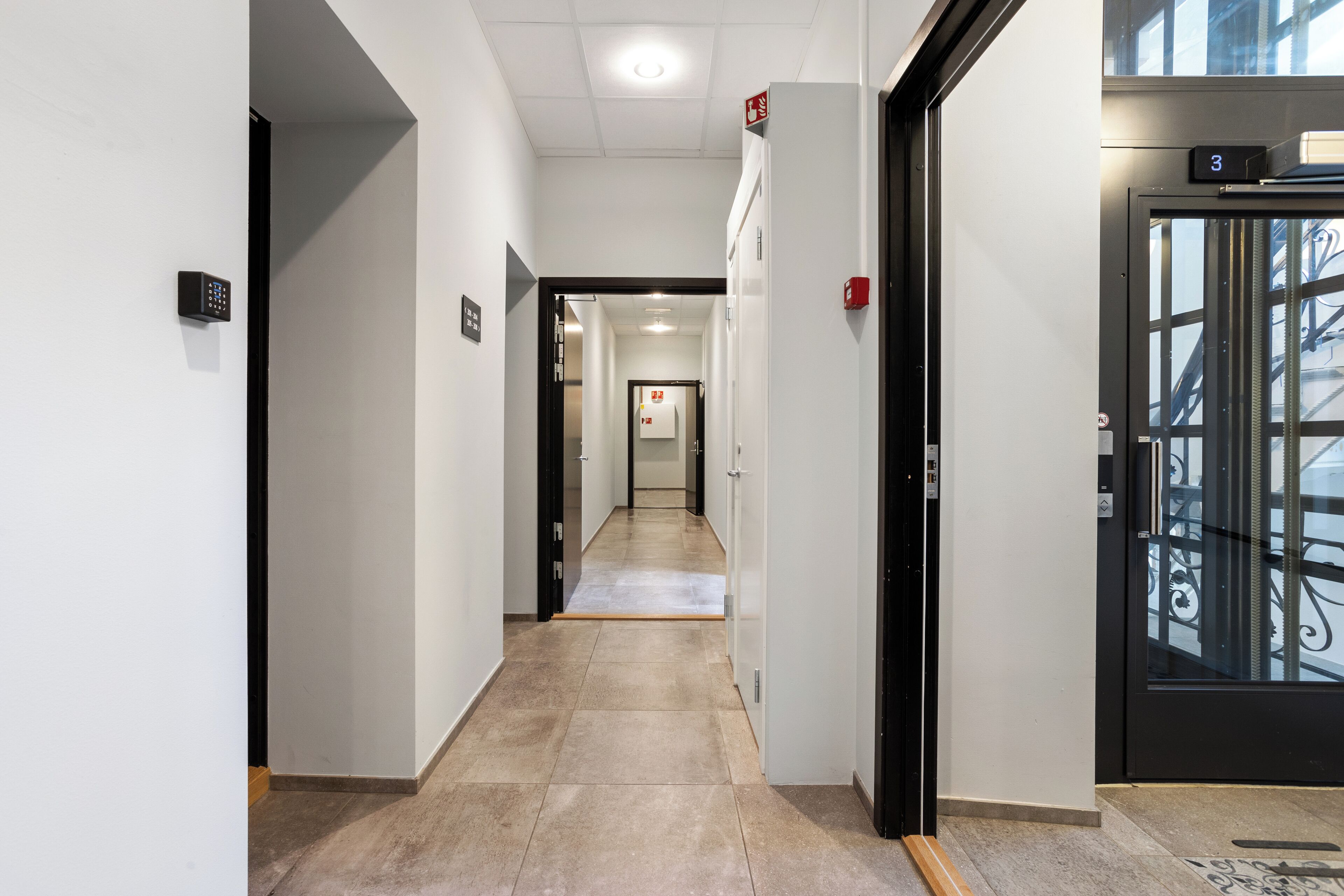 A modern hallway with a tiled floor, a black door with a number sign, and a glass elevator door.