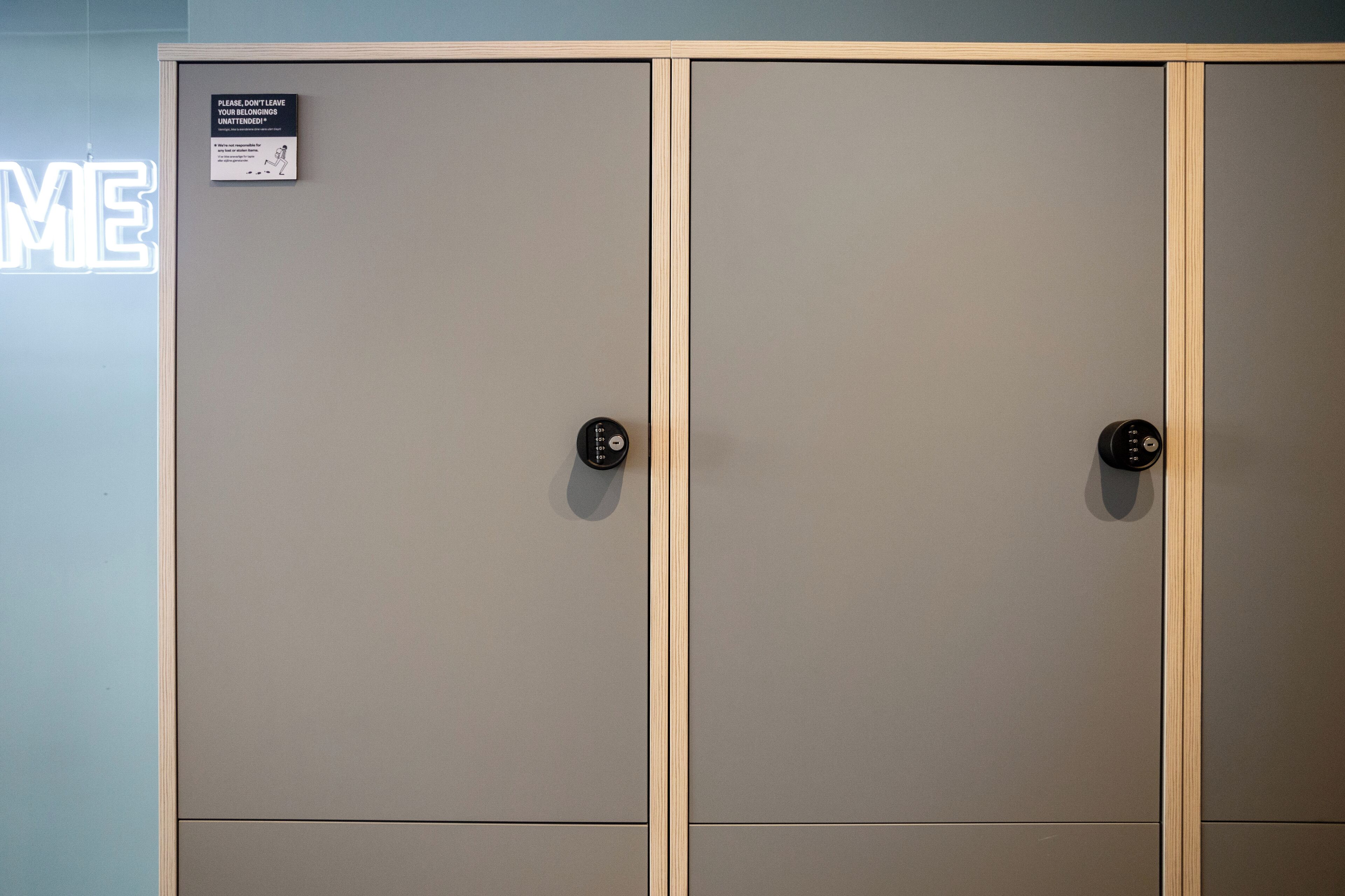 A set of grey lockers with a wooden frame, a sign instructing not to leave belongings unattended, and a neon sign with the word "ME" in the background.