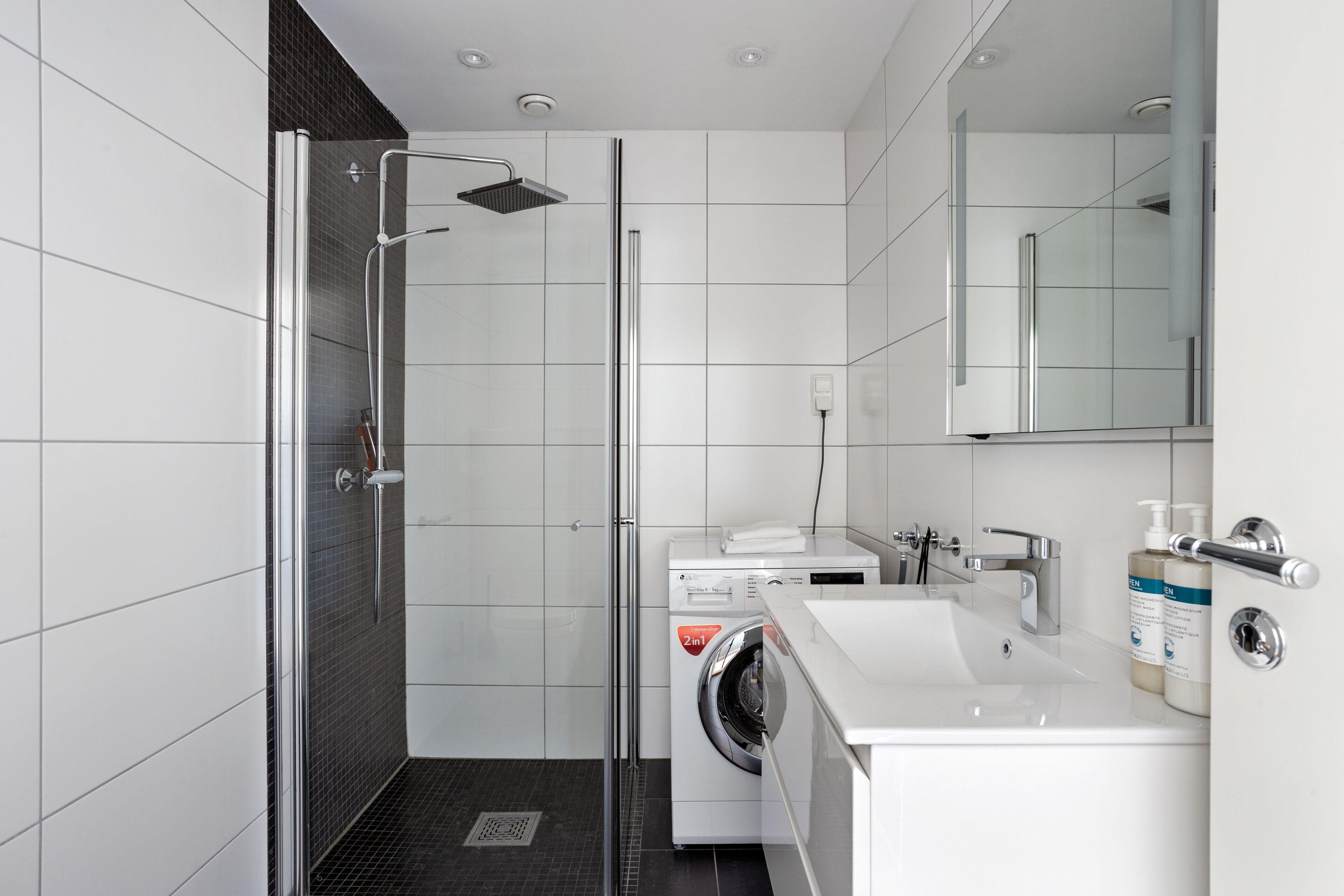 A modern bathroom with a glass shower enclosure, a built-in washing machine, and a sink with a mirror above it.