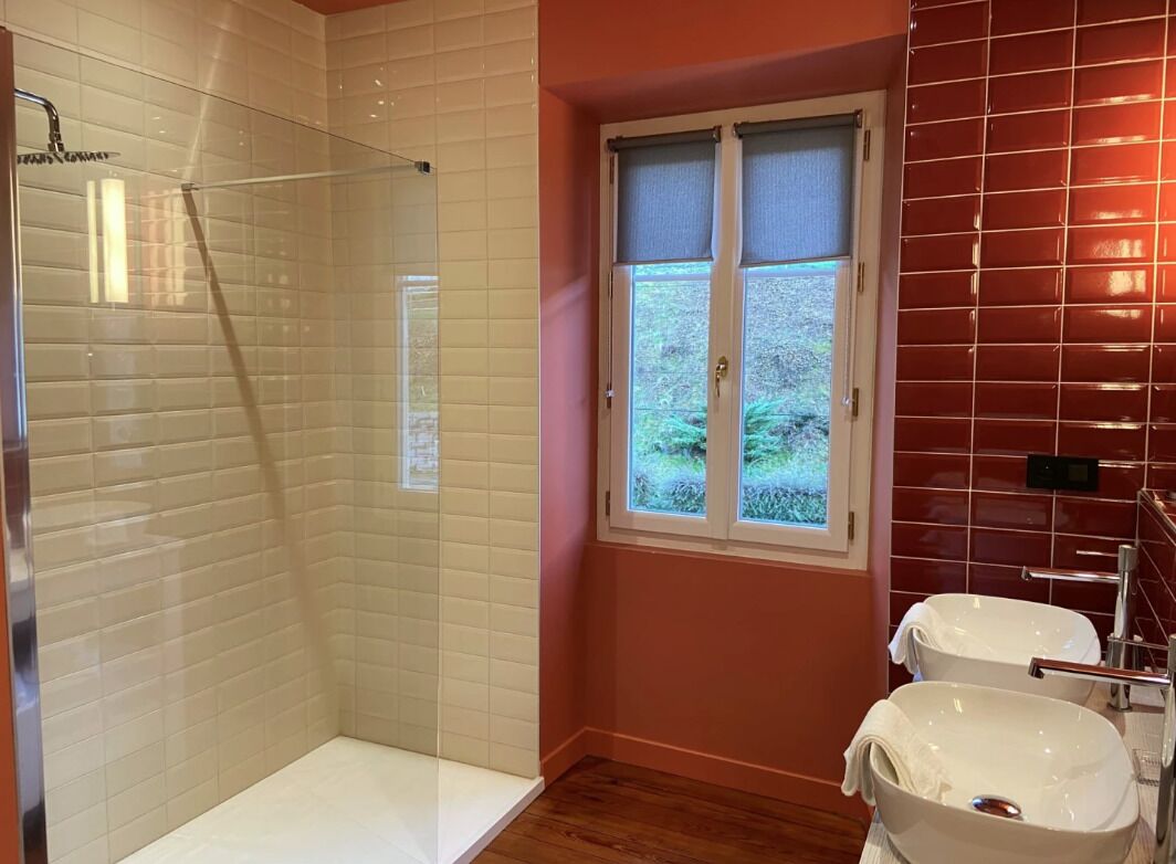 Un baño con cabina de ducha de vidrio, una ventana con vistas a la vegetación y dos lavamanos blancos.