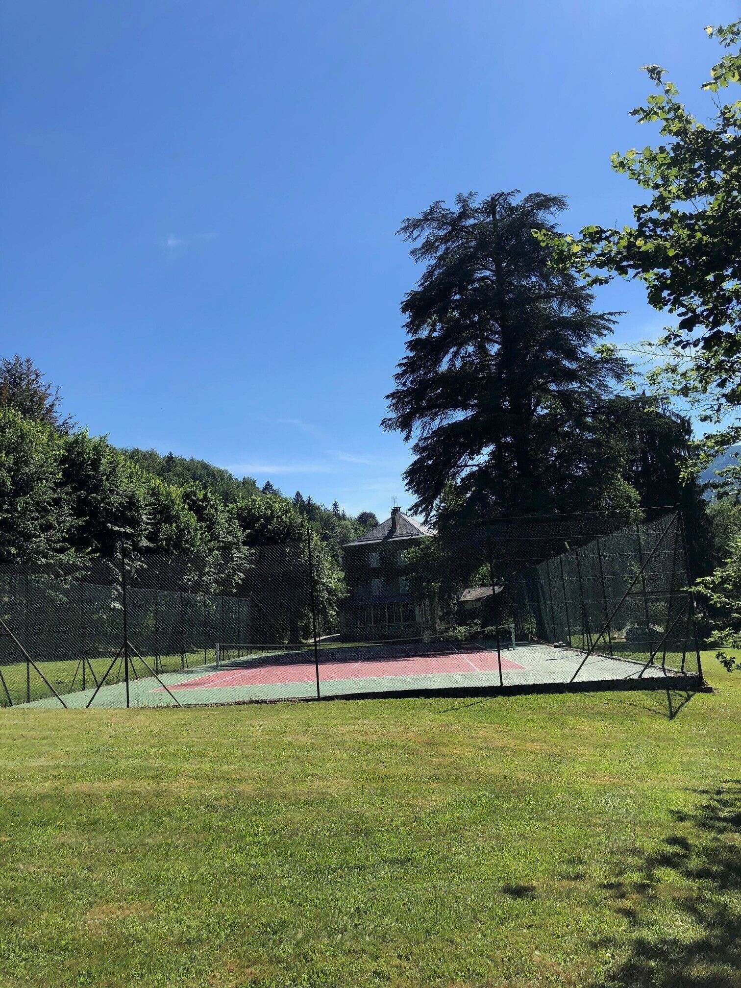 A tennis court surrounded by a fence, with a building in the background and trees around.
