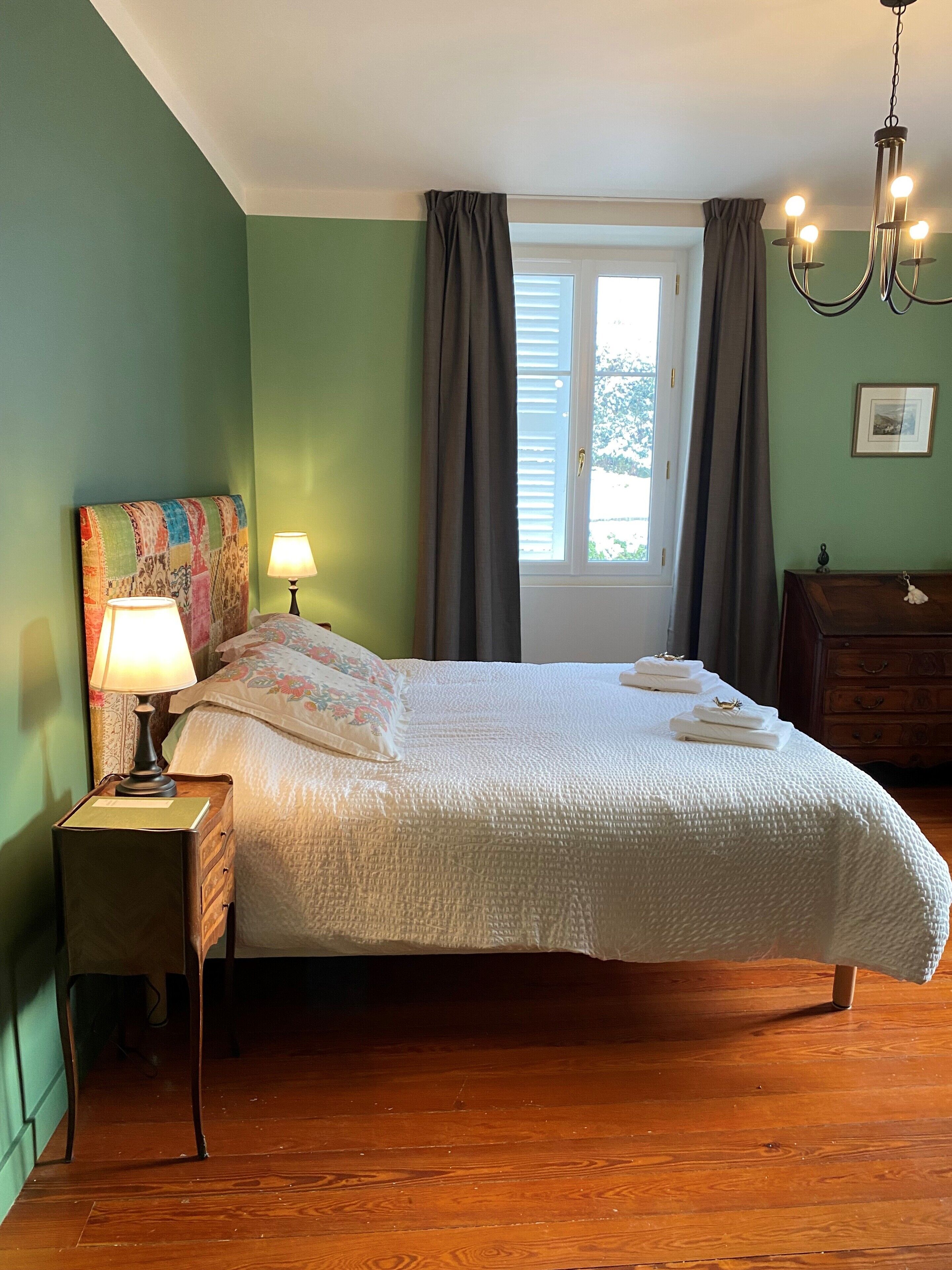 Un dormitorio con una cama, una mesita de noche con lámpara, un aparador, una ventana con cortinas y un candelabro.