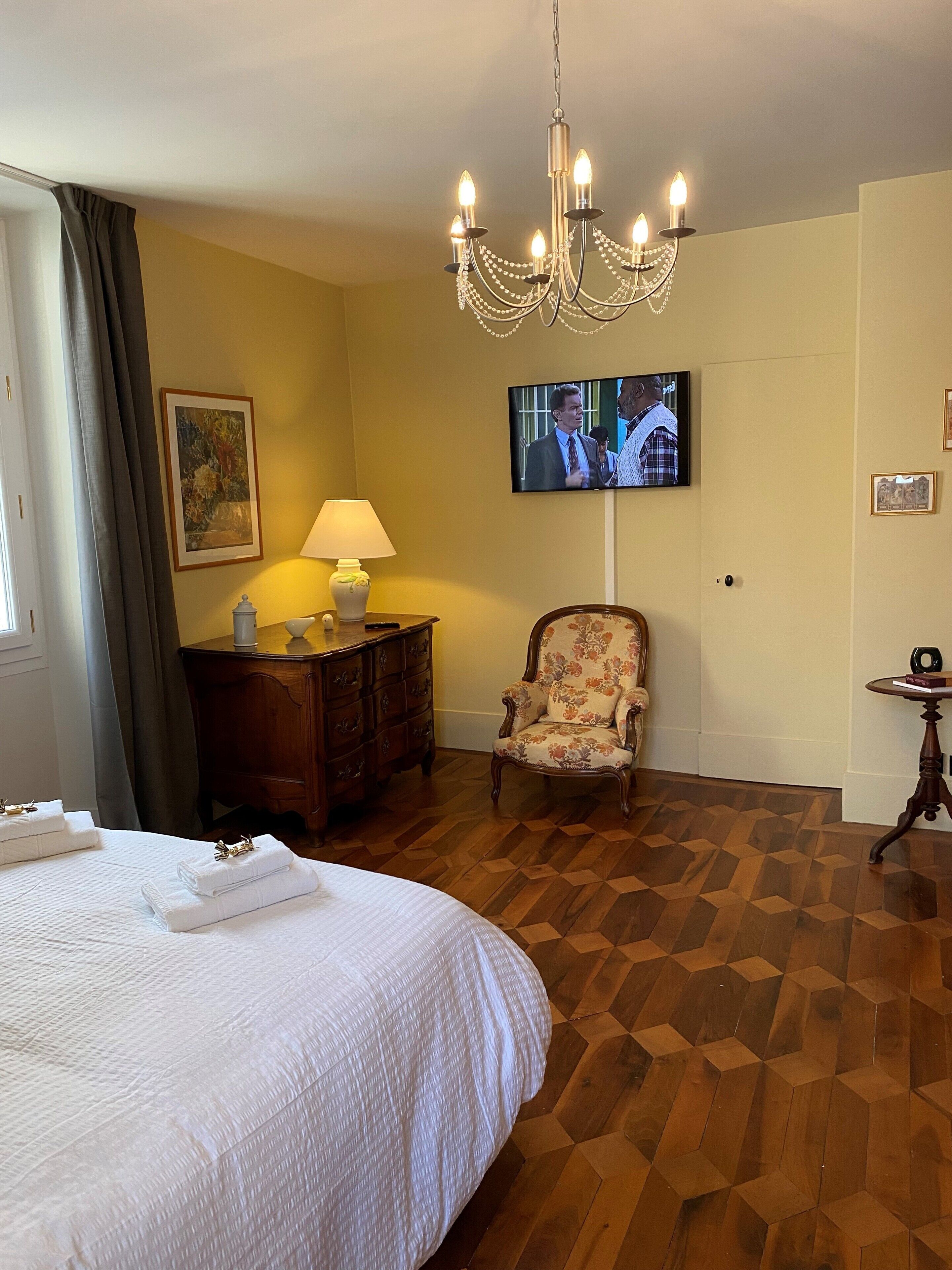 A hotel room with a large bed, a wooden floor, a television, a chair, and a desk with a lamp.