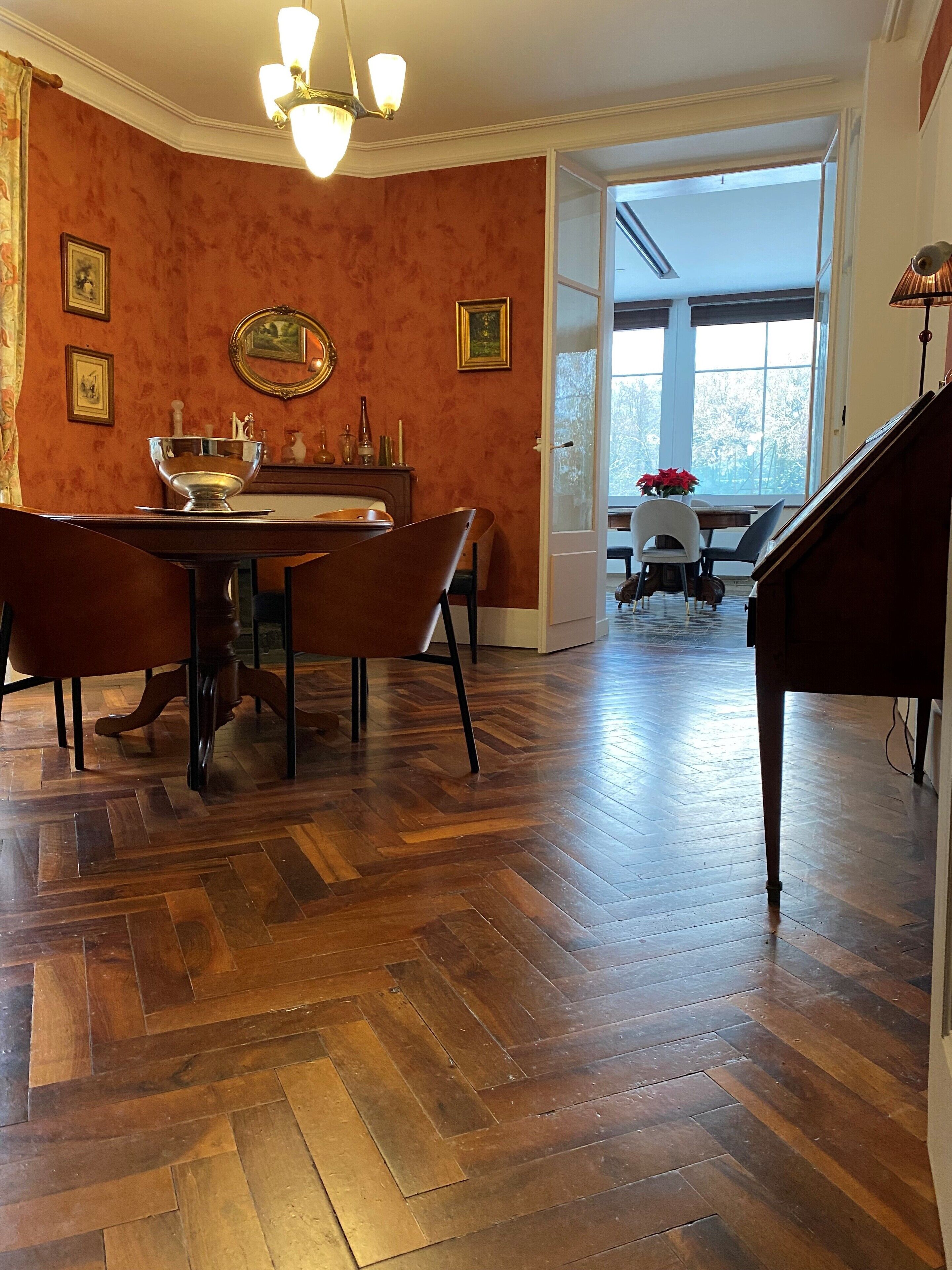 A dining area with a wooden herringbone floor, a dining table with chairs, a piano, and a large window providing natural light.