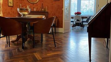 A dining area with a wooden herringbone floor, a dining table with chairs, a piano, and a large window providing natural light.