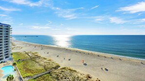 En strand med en lång pir som sträcker sig ut i havet, ett poolområde med solstolar och en byggnad med flera våningar.
