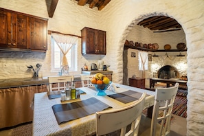 A rustic kitchen with stone walls, wooden cabinets, a stone fireplace, and a dining table set with placemats and a fruit bowl.