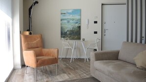 Un salon avec un canapé, un fauteuil et une petite table ronde sur laquelle se trouve un saladier rempli de fruits. Un tableau est accroché au mur, et une porte est visible au fond.
