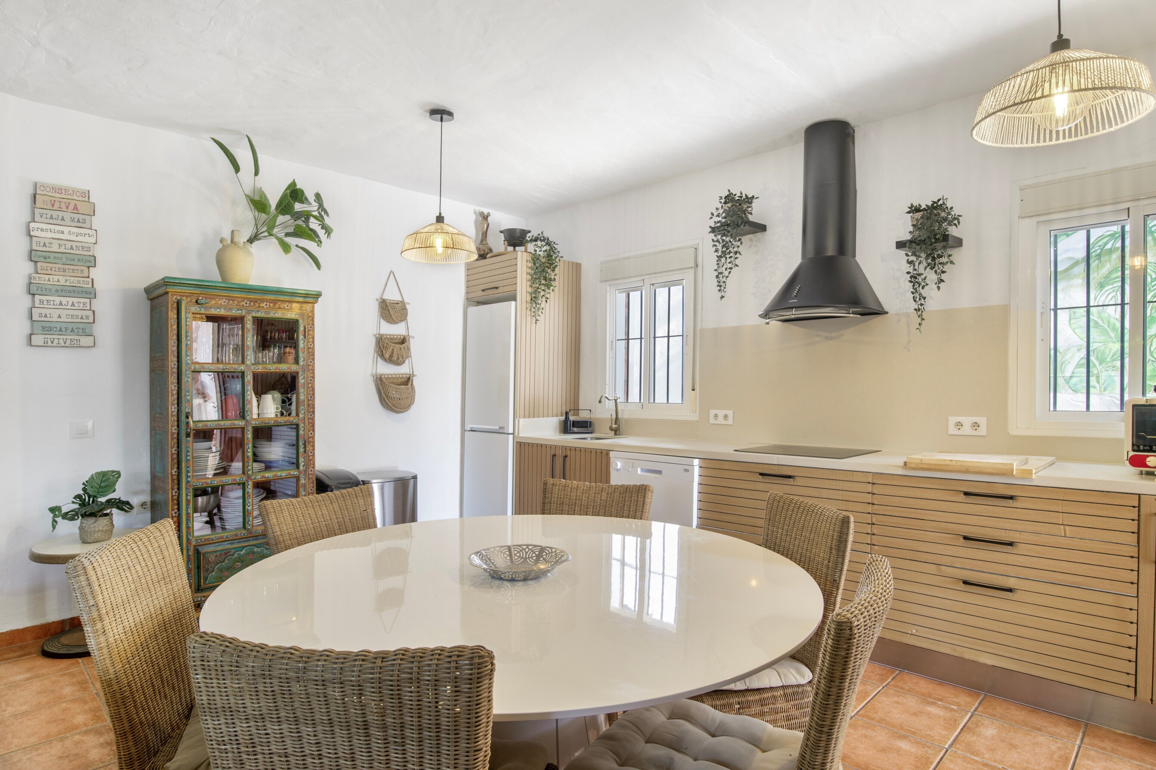 A kitchen with a round dining table, wicker chairs, a built-in bookshelf, and hanging plants.