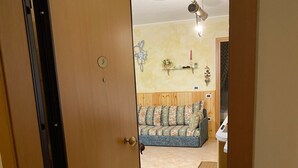 Une pièce avec une porte en bois, un sol carrelé, un canapé, une étagère fixée au mur ornée d’objets décoratifs et un ventilateur de plafond.