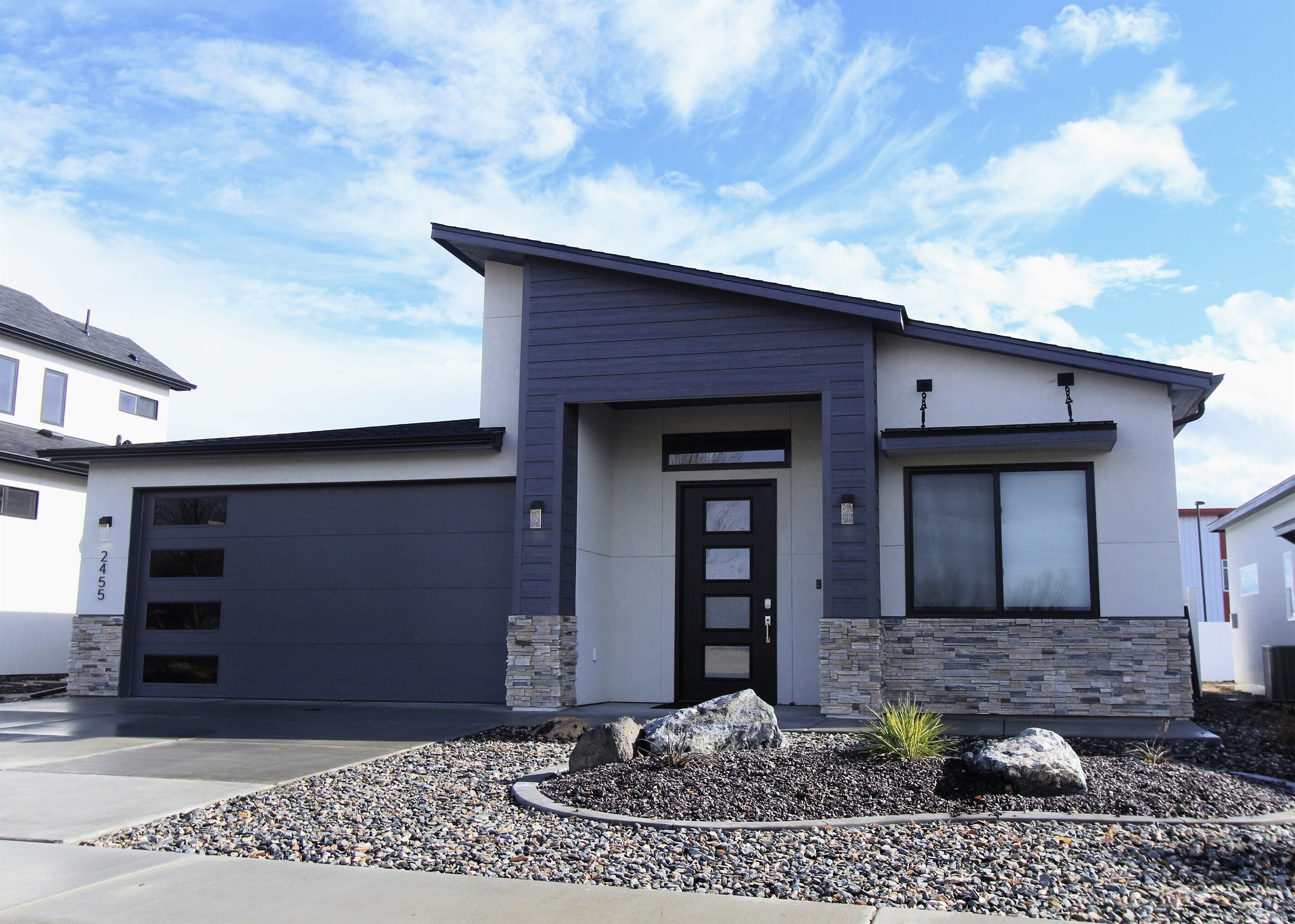 Een modern tweegezinshuis met garage, een geaccentueerde stenen muur en een aangelegde voortuin met rotsen en beplanting.