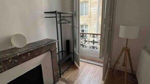 Ein schmales Zimmer mit Holzboden, Kamin, einem Metallregal und einem Balkon mit schmiedeeisernem Geländer.