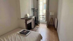 Ein schmales Zimmer mit Bett, einem kleinen Tisch, einem Heizkörper und einem Fenster.
