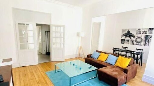 Ein Wohnzimmer mit einem Glastisch, einem braunen Sofa mit bunten Kissen, einem blauen Teppich und einem Essbereich mit Tisch und Stühlen.