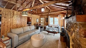 A rustic cabin interior with a wooden ceiling, a fireplace, a sofa, a coffee table, and a dining table.