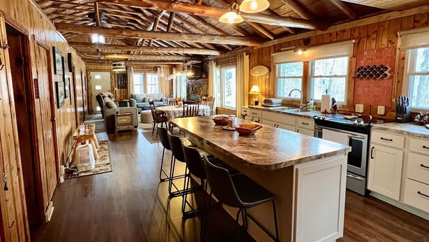 A spacious kitchen with a large island, wooden ceiling beams, and a view of the outdoors through large windows.