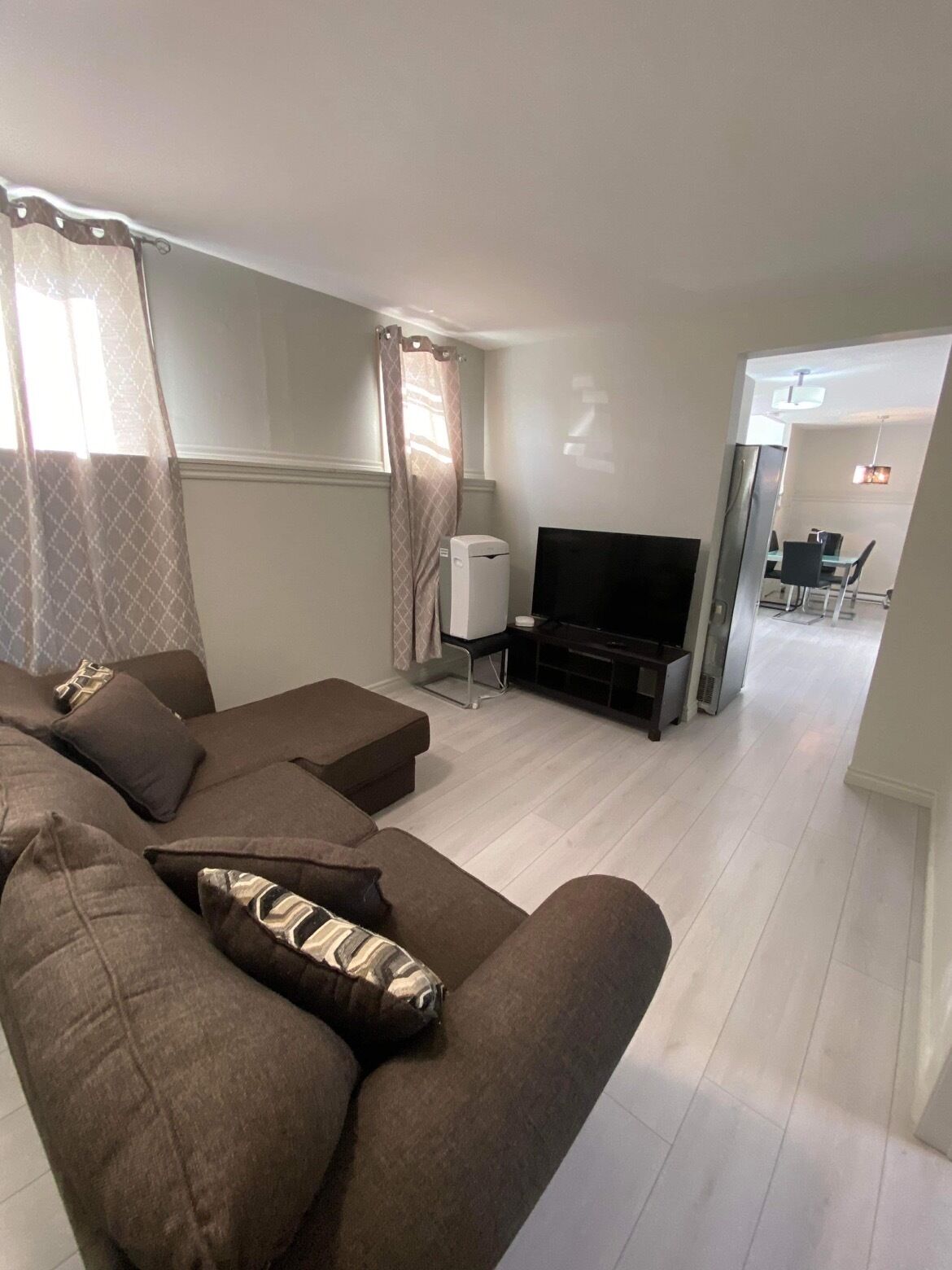 A modern living room with a sectional sofa, a flat-screen TV, and a dining area visible through an open doorway.