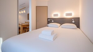 A modern hotel room with a large bed, bedside lamps, and a view into the dining area.