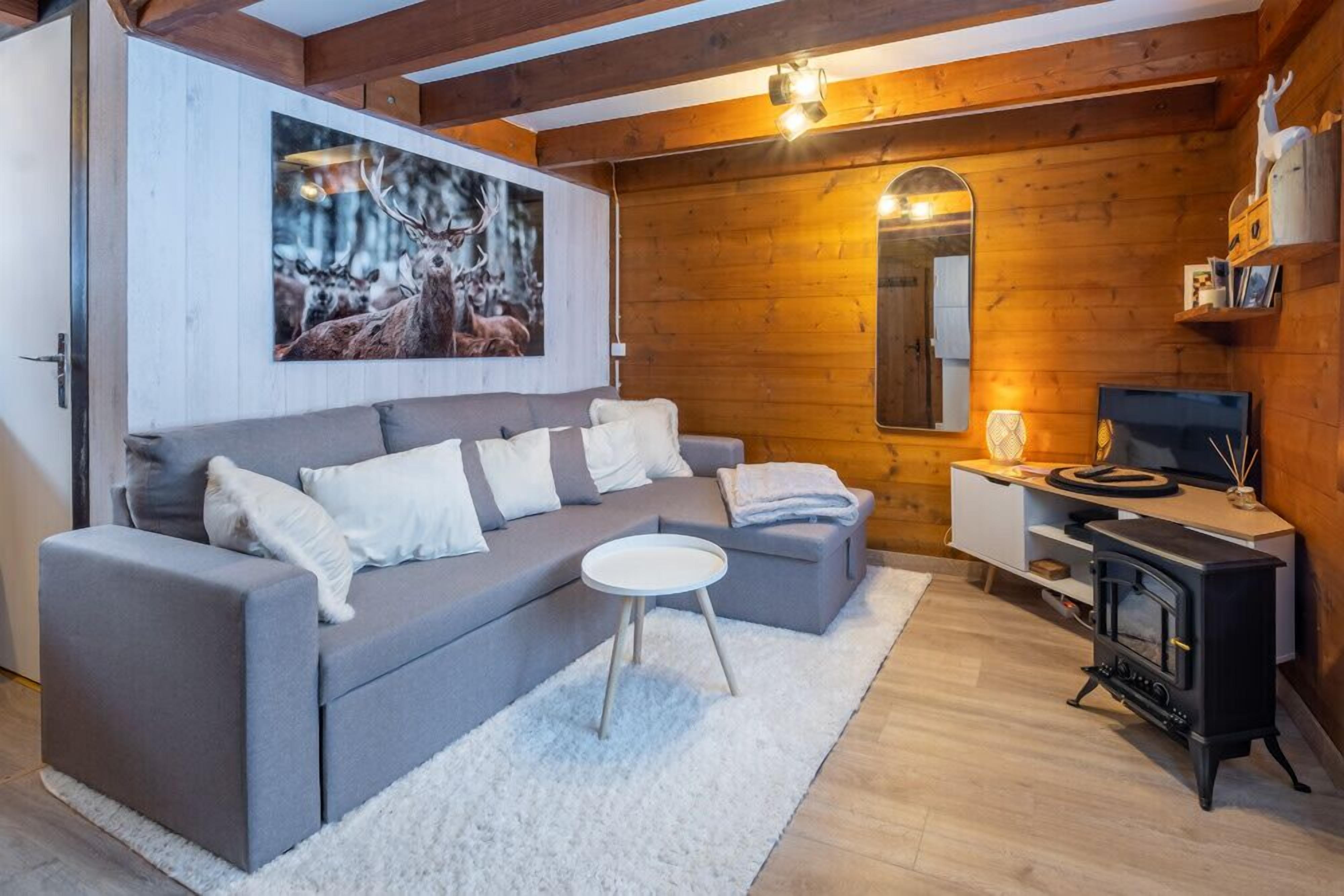 Ein gemütliches Wohnzimmer mit grauem Sofa, einem kleinen weißen Tisch, einer Holzwand mit Spiegel und einem schwarzen Kaminofen.